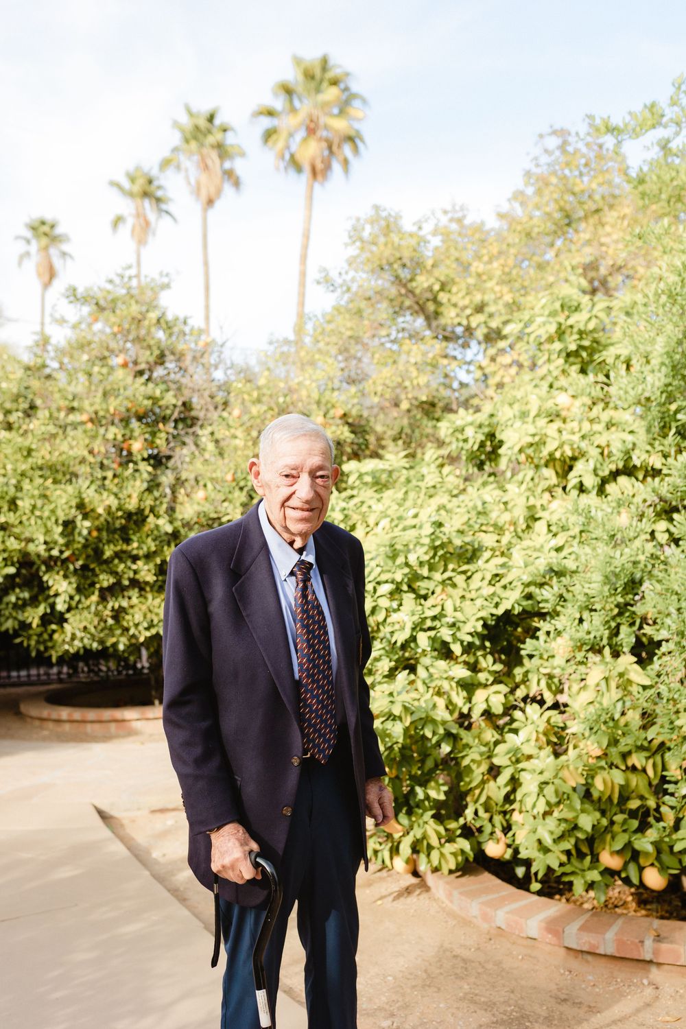 An elderly person in a dark suit and tie stands outdoors with palm trees and greenery in the background.
