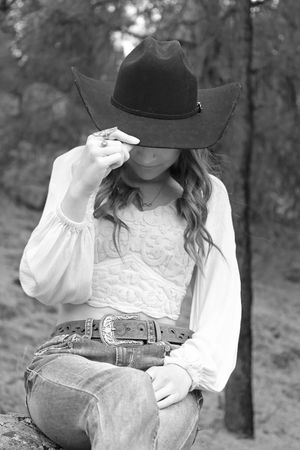 Black and white portrait of young woman in white lace top tipping dark cowboy hat outdoors.
