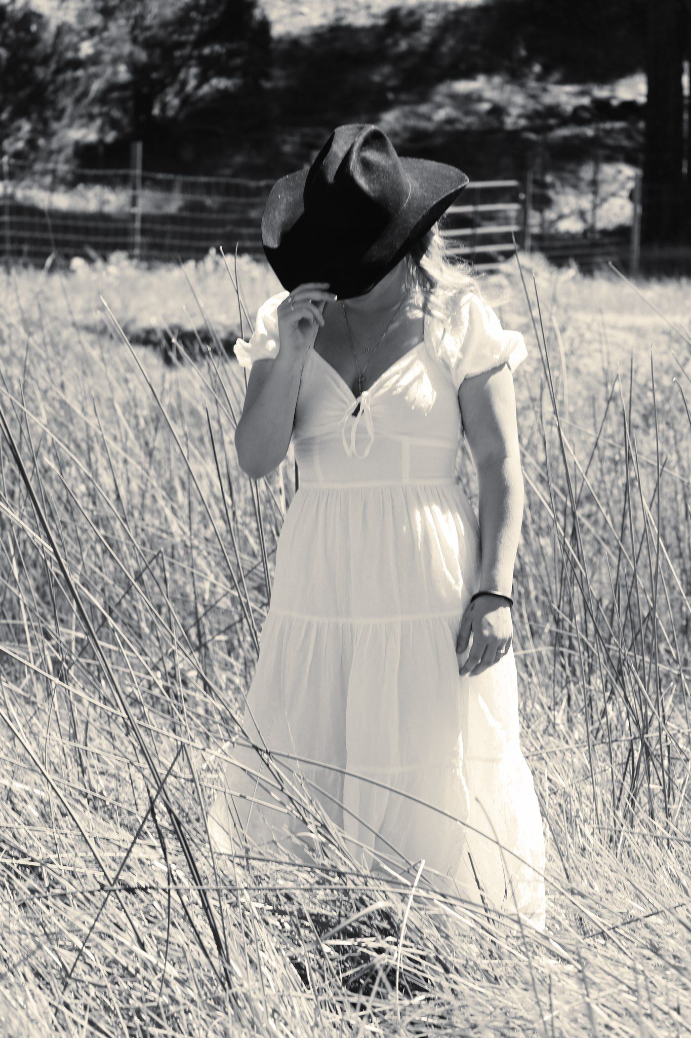 Black and white photo of woman in white dress tipping cowboy hat in tall grass field.