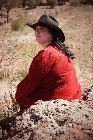 Woman in red shirt and black cowboy hat sitting on rocks in a rural outdoor setting.