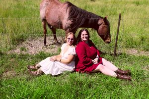 Two women in red and white outfits sitting on grass with a brown horse grazing behind them.