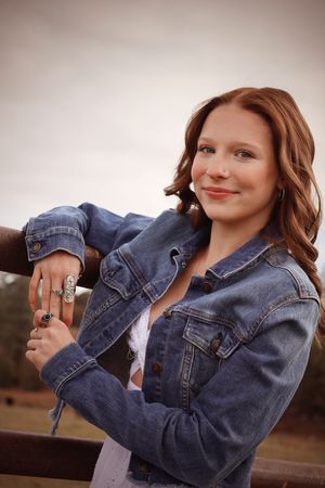 Young woman with red hair in a denim jacket leaning on a fence rail outdoors.