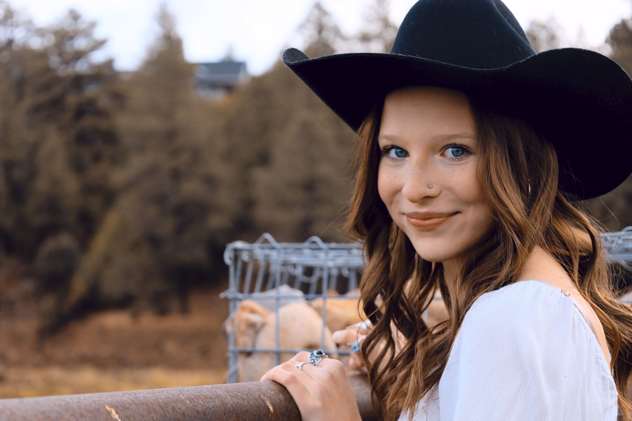 Young woman with long hair wearing a black cowboy hat smiling outdoors near a fence.