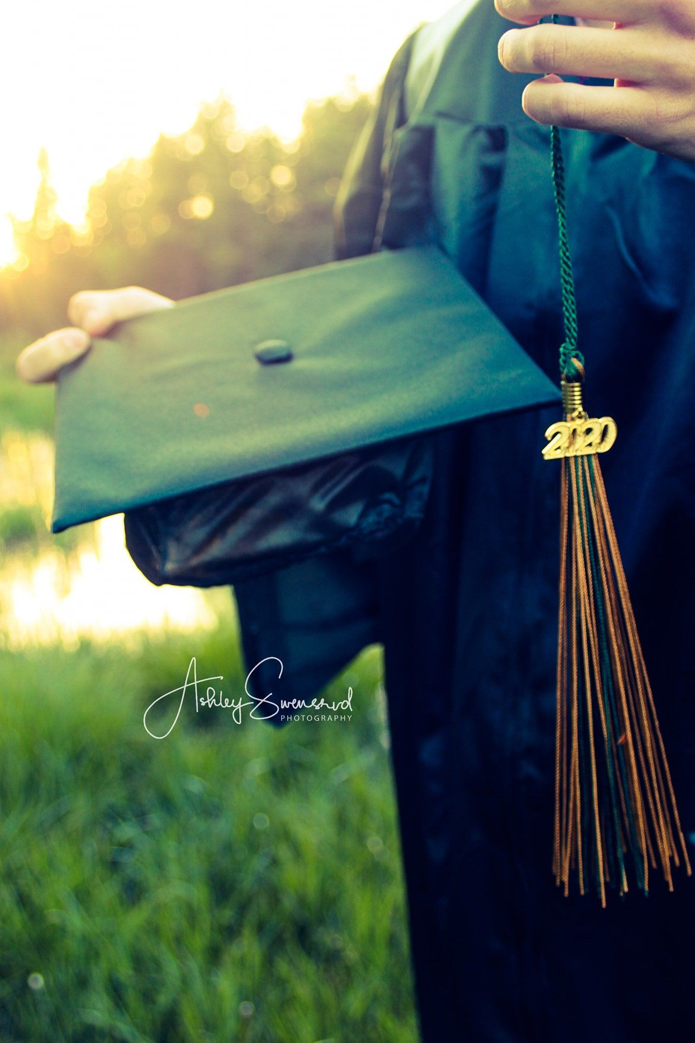 Graduate holding green 2020 mortarboard cap with gold tassel in sunlit outdoor setting.