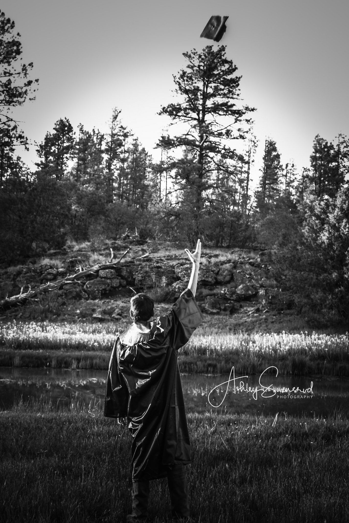 Black and white photo of graduate tossing cap into air near forest pond at sunset.