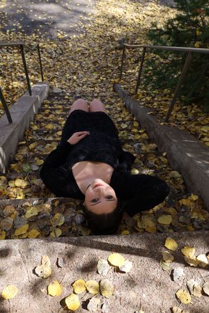 Woman in black dress lying on stone steps surrounded by fallen yellow autumn leaves.
