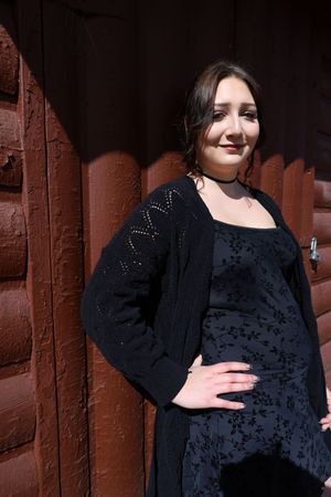 Young woman in black floral dress and cardigan posing against a rustic red log wall.