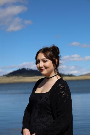 Young woman in black outfit standing near a lake with blue sky and mountain in background.