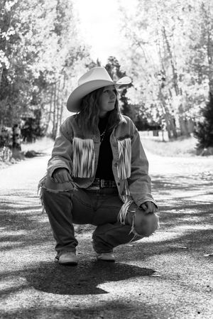 Black and white photo of a young woman in cowboy hat and fringed denim jacket crouching on a tree-lined path.