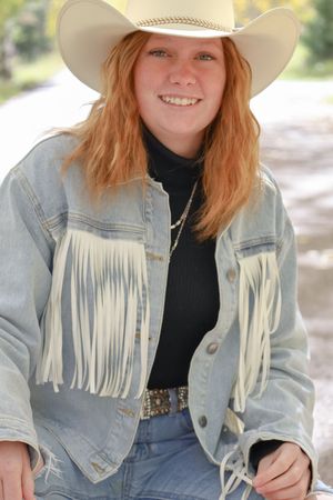Smiling redhead teen in white cowboy hat and fringed denim jacket with a black turtleneck and western belt.