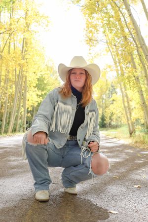 Redhead young woman in cowboy hat and fringed denim jacket crouching on a road surrounded by golden aspen trees.