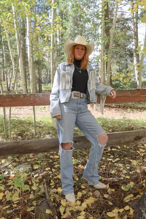 Young redhead woman in cowboy hat and fringed denim jacket leaning on a wooden fence in an autumn forest.
