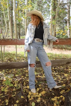 Redhead woman in white cowboy hat and fringed denim jacket posing by a rustic wooden fence in fall forest.