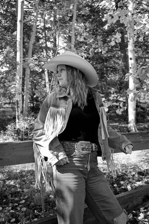 Black and white photo of a young woman in cowboy hat and fringed jacket leaning on a wooden fence outdoors.