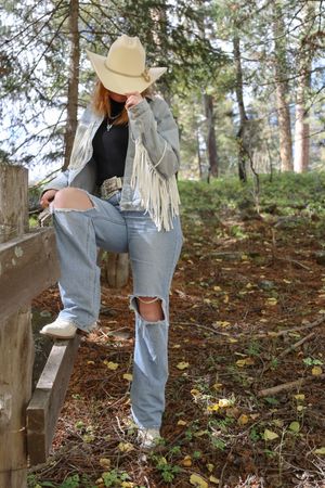 Young woman in cowboy hat and fringed jacket sitting on a wooden fence rail in a forest setting.