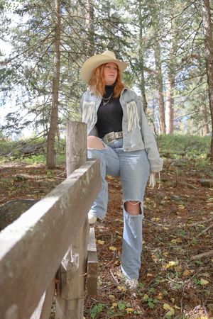 Young woman in fringe denim jacket, cowboy hat, and ripped jeans posing by rustic wooden fence in autumn forest.
