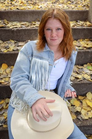 Redhead woman in fringe denim jacket holding cowboy hat while sitting on stone steps covered in yellow fall leaves.