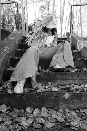 Black and white photo of woman in fringe jacket and wide-leg jeans sitting on leaf-covered outdoor stone steps.