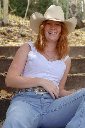 Smiling redhead woman in white tank top, cowboy hat, and embellished belt sitting on stone steps in autumn setting.
