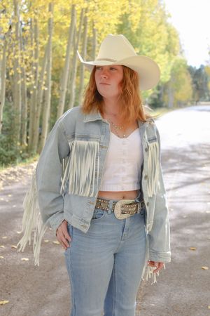 Redhead woman in cream cowboy hat and fringe denim jacket walking on rural road lined with golden autumn trees.