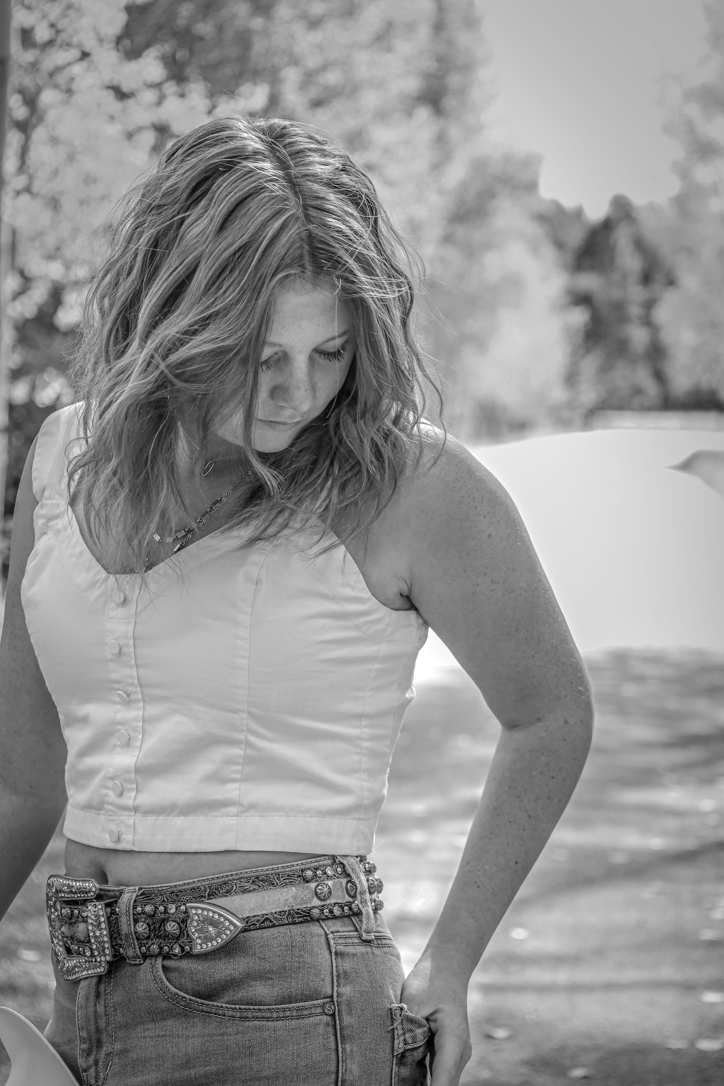 Black and white portrait of woman in white crop top and studded western belt with wind-blown hair looking down.