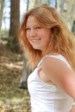 Smiling redhead woman in white tank top outdoors in sunny autumn woods.