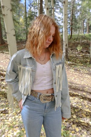 Redhead woman in fringed denim jacket and jeans stands in fall aspen forest.