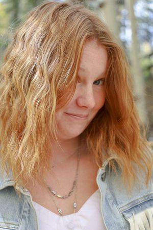 Close-up of redhead woman in denim jacket with layered necklaces in forest.