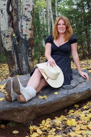 Smiling redhead woman in black dress holds cowboy hat seated on rock in autumn.