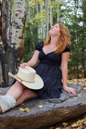 Redhead woman in black dress and cowboy boots gazes up while seated on forest rock.