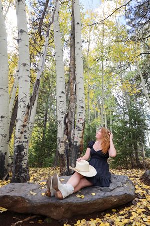 Redhead woman in black dress with cowboy hat sits on rock among fall aspen trees.