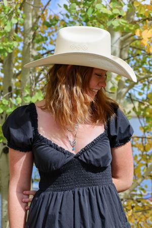 Redhead woman in black dress and white cowboy hat smiles in fall aspen grove.