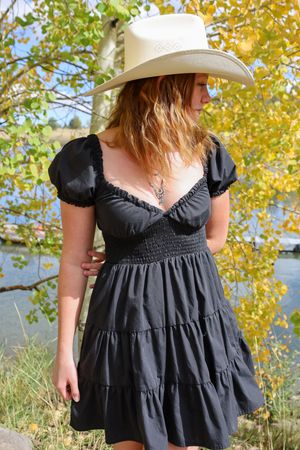 Redhead woman in black mini dress and white cowboy hat near autumn riverside trees.