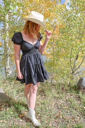 Redhead woman in black mini dress tips white cowboy hat in autumn aspen setting.