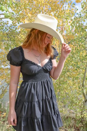 Young redhead woman in white cowboy hat and black tiered dress among fall foliage.