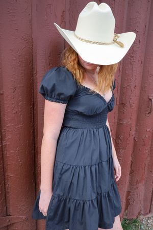 Redhead woman in white cowboy hat and black dress posing against a rustic red barn wall.