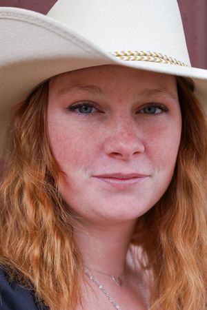 Close-up portrait of a freckled redhead woman wearing a white cowboy hat with gold trim.