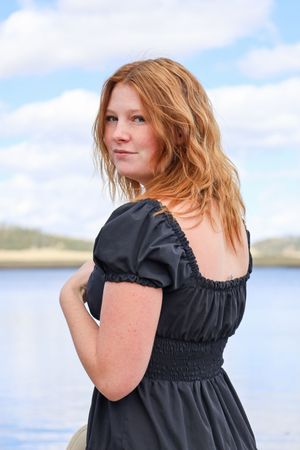 Young redhead woman in black smocked dress posing outdoors near a serene lake.