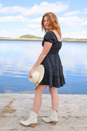 Redhead woman in black dress holding white cowboy hat standing on a wooden dock by a lake.