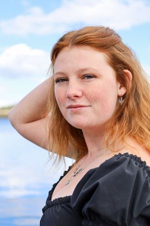 Portrait of a smiling redhead woman in a black dress with a lake and blue sky behind her.