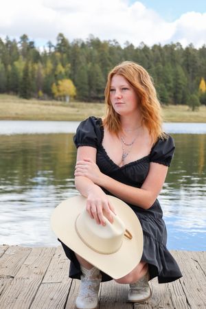 Redhead woman in black dress crouching on a dock holding a white cowboy hat by a lake.