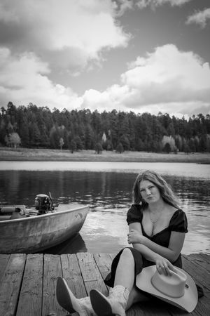 Black and white photo of woman in black dress with cowboy hat sitting on dock beside lake and forest.