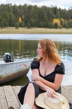 Redhead woman in black dress holding cowboy hat sits on wooden dock by lake with boat and autumn forest.