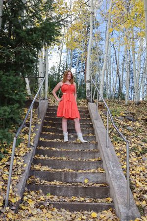 Smiling redhead woman in orange dress and white boots stands on stone stairs covered in autumn leaves.