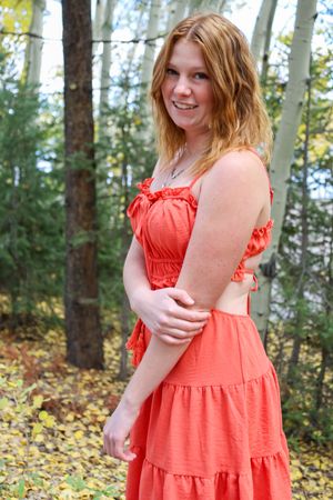 Smiling redhead woman in orange ruffled dress poses among aspen trees with yellow autumn leaves on ground.