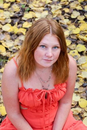 Close-up portrait of blue-eyed redhead woman in orange dress sitting among vibrant yellow fallen leaves.