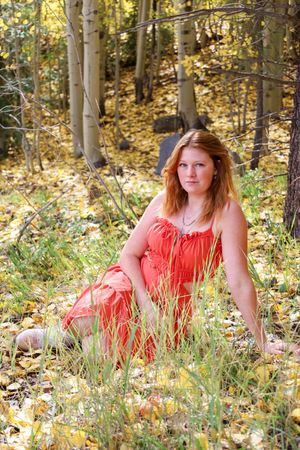 Redhead woman in long orange dress sits in tall grass among aspen trees with golden autumn foliage.