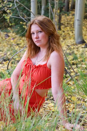 Young redhead woman in red dress sitting in autumn forest grass.
