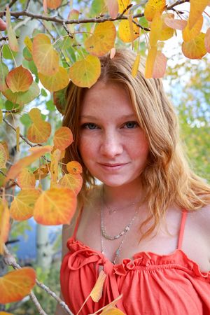 Smiling redhead teen with freckles in coral top surrounded by colorful autumn aspen leaves.