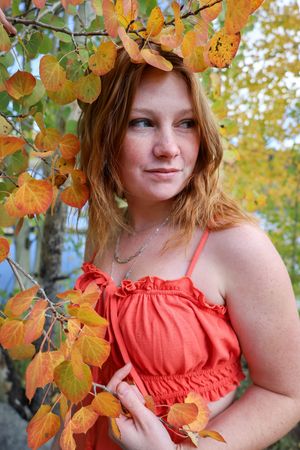 Young redhead woman in orange sundress posing among vibrant orange and yellow fall aspen leaves.